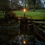 Large railway style cast iron pedestal post light in black, with a round hooped shade and glowing bulb, set on a lawn beside stone steps and water.