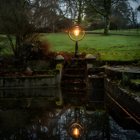 Large railway style cast iron pedestal post light in black, with a round hooped shade and glowing bulb, set on a lawn beside stone steps and water.