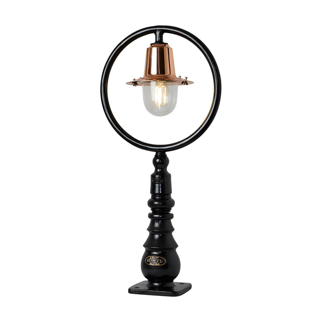 Small railway-style cast iron pedestal post light with a black circular hoop frame, copper spun shade over a clear glass dome, ribbed black base, on a plain white background.