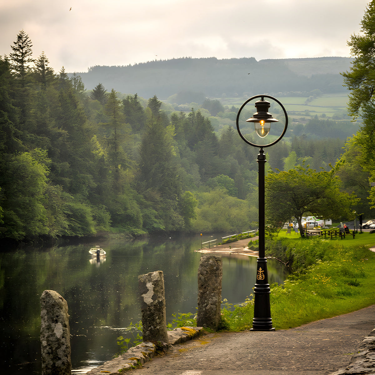 Railway style cast iron lamp post in black, 3.65m tall, with a spun shade under a hoop and clear glass-effect dome, shown lit beside a riverside path.