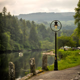 Railway style cast iron lamp post in black, 3.65m tall, with a spun shade under a hoop and clear glass-effect dome, shown lit beside a riverside path.
