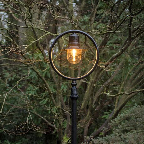 Railway Style cast iron lamp post in black, 2.73m tall, with a lit lantern head inside a circular hoop frame, set outdoors against bare and green trees.
