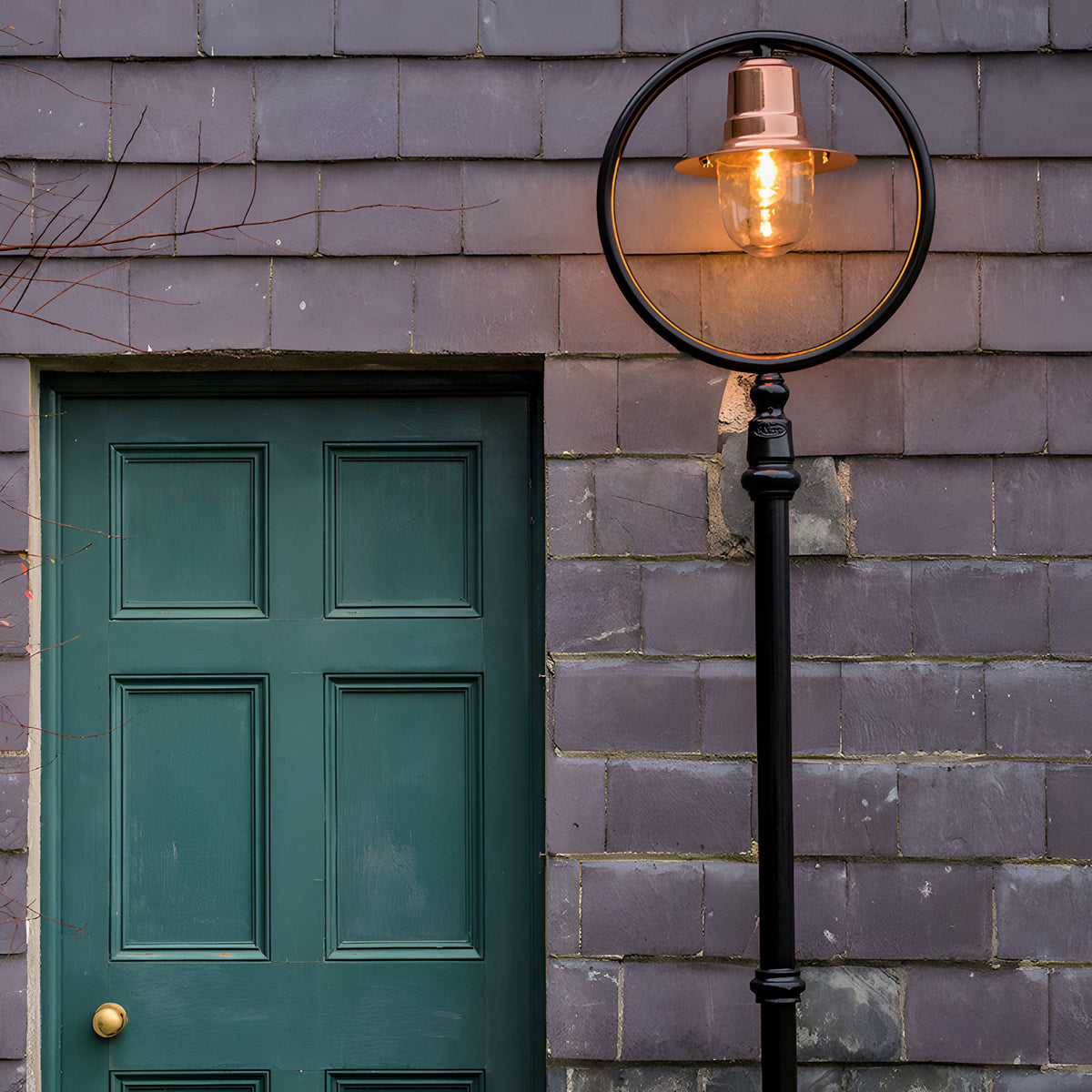 Railway Style cast iron lamp post, 2.43m, black frame with circular hoop, copper spun shade over a lit glass dome, mounted beside a green panelled door on a grey tiled wall.