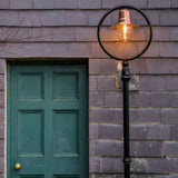 Railway Style cast iron lamp post, 2.43m, black frame with circular hoop, copper spun shade over a lit glass dome, mounted beside a green panelled door on a grey tiled wall.