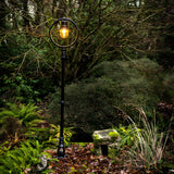 Railway Style Cast Iron Lamp Post, 2.43m, in black, with a circular hoop frame around a lit globe shade atop a fluted post, set in a garden path.
