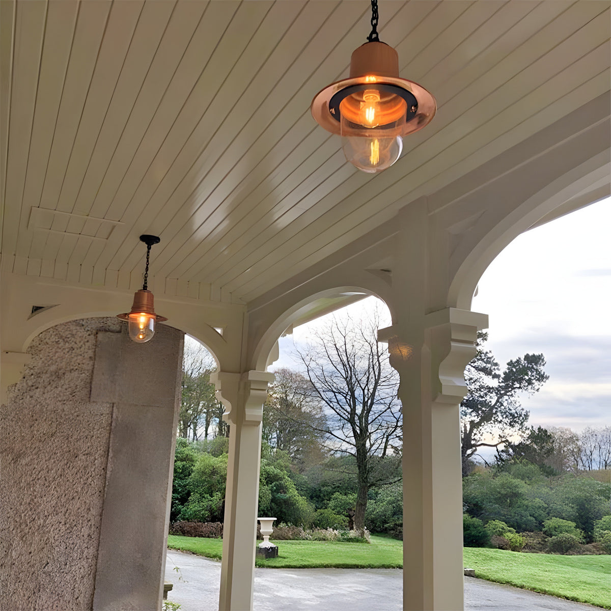 Medium Railway Style Hanging Lantern on a chain with copper shade and clear glass dome, illuminated on a covered porch ceiling overlooking a garden.