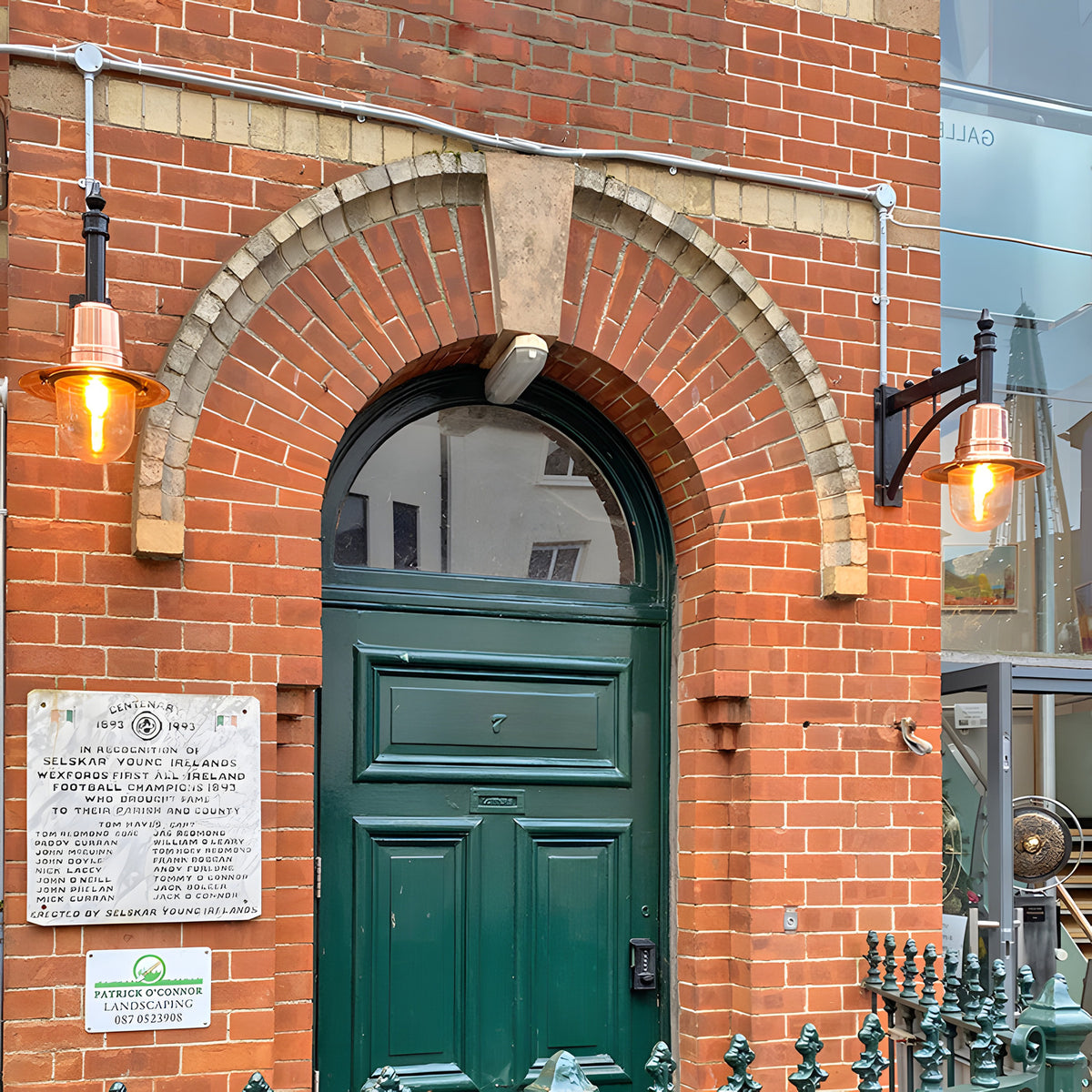 Medium railway style wall lantern with copper shade on a cast arm, mounted either side of an arched green front door on a red brick building.