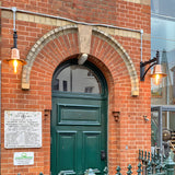 Medium railway style wall lantern with copper shade on a cast arm, mounted either side of an arched green front door on a red brick building.