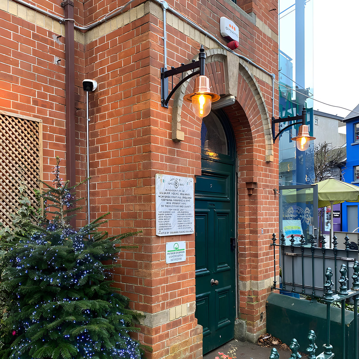 Medium railway style wall lantern with copper shade on a black cast arm, mounted beside an arched doorway on a brick building exterior.