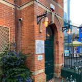 Medium railway style wall lantern with copper shade on a black cast arm, mounted beside an arched doorway on a brick building exterior.