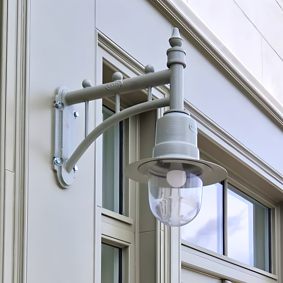 Medium Railway Style Wall Lantern on a cast arm, grey traditional outdoor wall light with domed clear glass shade, mounted on a building exterior beside windows.