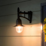 Small Railway style black wall lantern on a trussed cast arm with a clear glass dome, mounted outdoors on wooden cladding, lit at dusk.