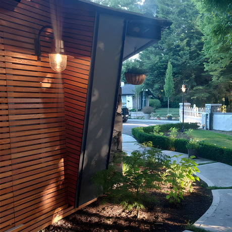Small vintage wall lantern with copper teardrop shade on a black goose neck bracket, mounted on cedar slat cladding beside a garden pathway.