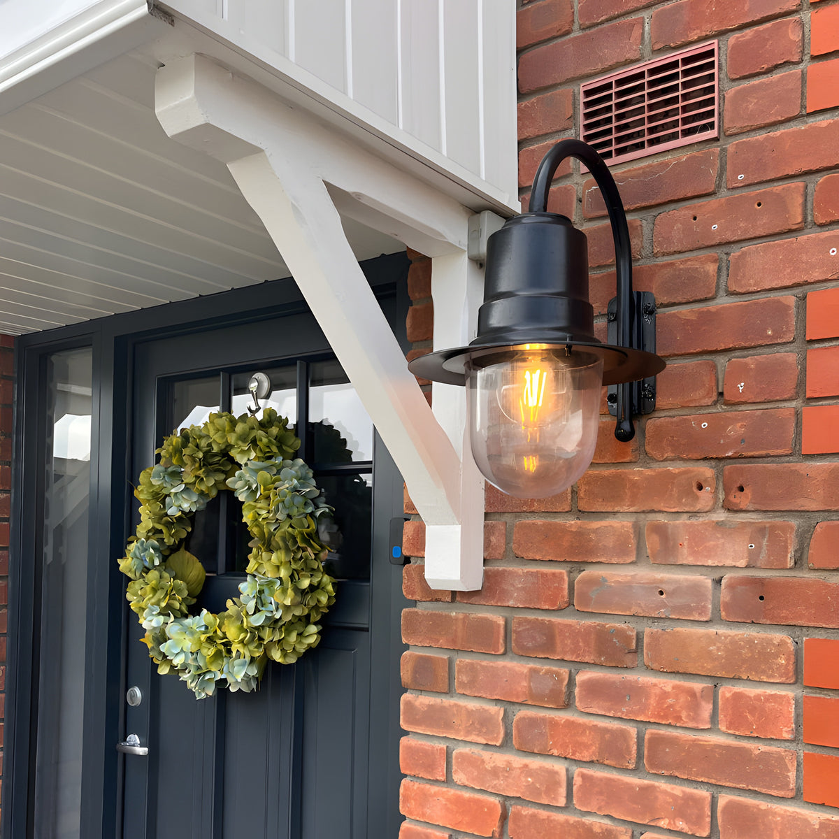 Small Vintage Wall Lantern on a black goose neck bracket, mounted on a brick wall beside a dark front door, with teardrop shade over a lit clear glass globe.