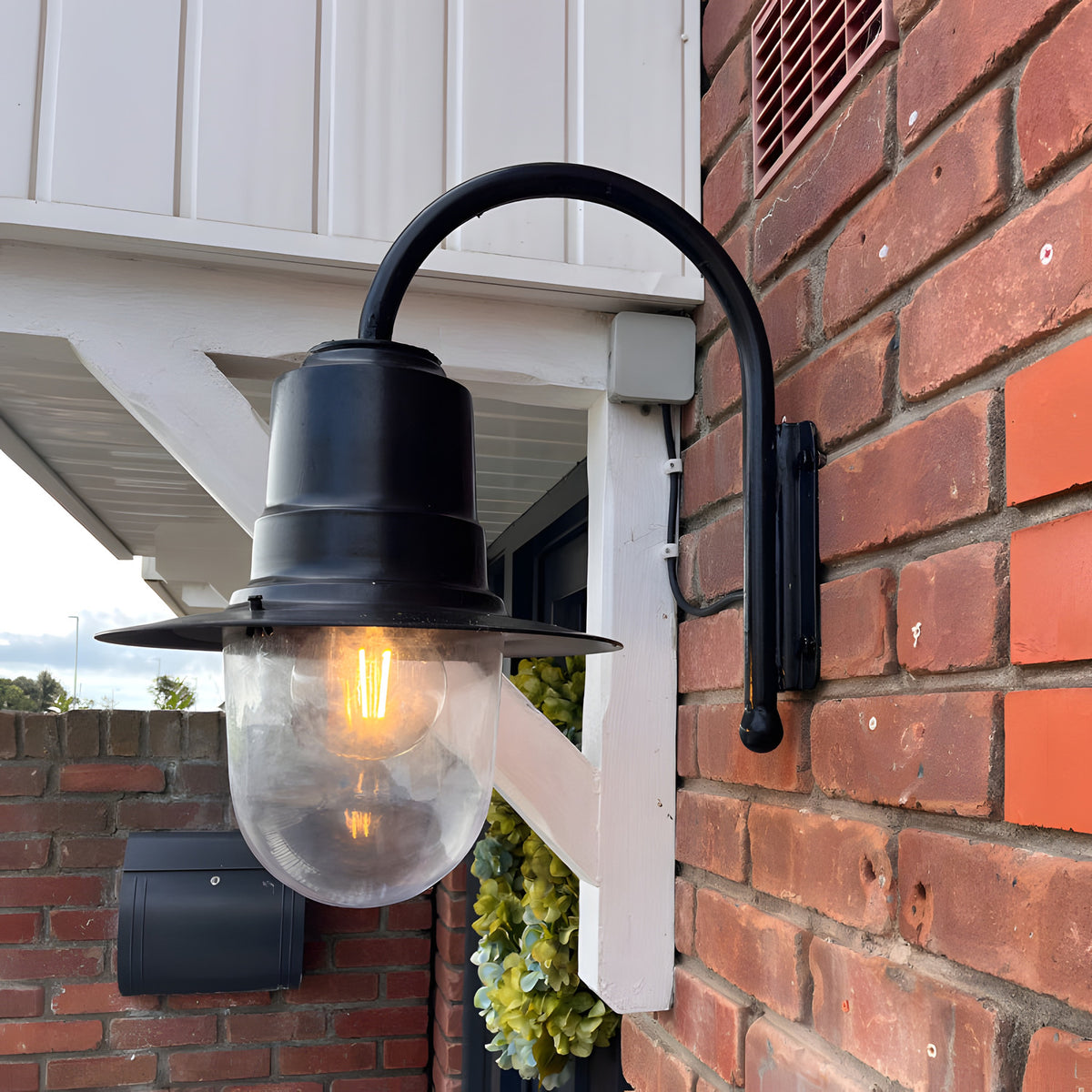 Small Vintage Wall Lantern on a black gooseneck bracket, mounted on a brick wall with a lit E27 bulb inside a clear polycarbonate shade.