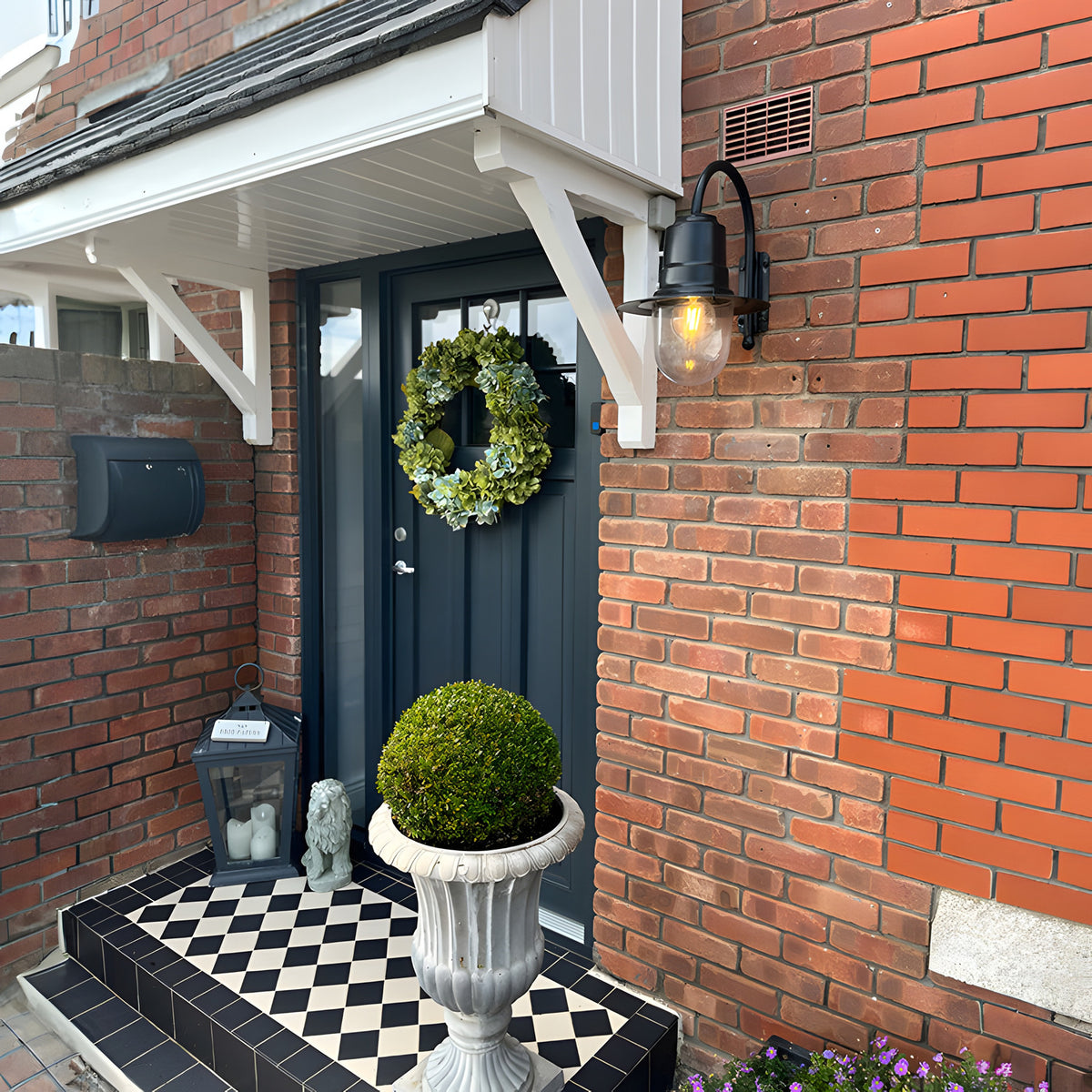 Small vintage wall lantern in black on a goose neck bracket, glass cylinder shade, lit bulb, mounted on a brick wall beside a dark front door.