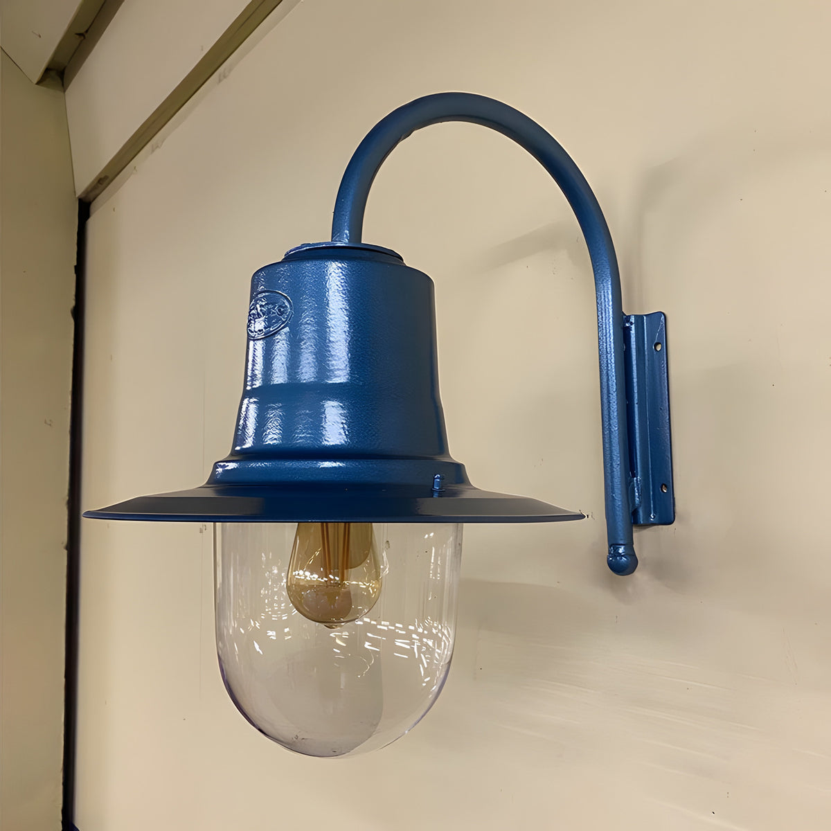 Small vintage wall lantern in blue on a goose neck bracket, with a teardrop shade over a clear polycarbonate cylinder and visible bulb, mounted on a plain wall.