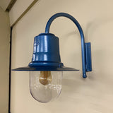 Small vintage wall lantern in blue on a goose neck bracket, with a teardrop shade over a clear polycarbonate cylinder and visible bulb, mounted on a plain wall.