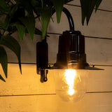 Small Vintage Wall Lantern on a black goose neck bracket, illuminated with a bell-shaped shade over a clear polycarbonate cylinder, mounted on a wooden panelled wall amid foliage.