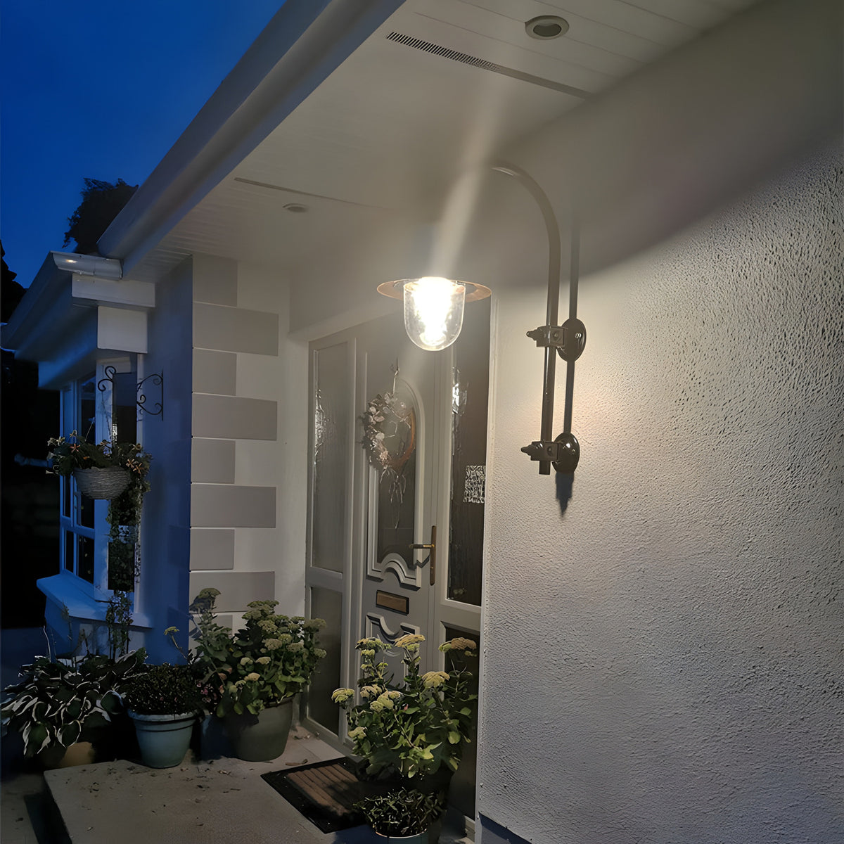 Large vintage wall lantern on a black goose neck bracket, illuminated beside a front door on a rendered house exterior at dusk.