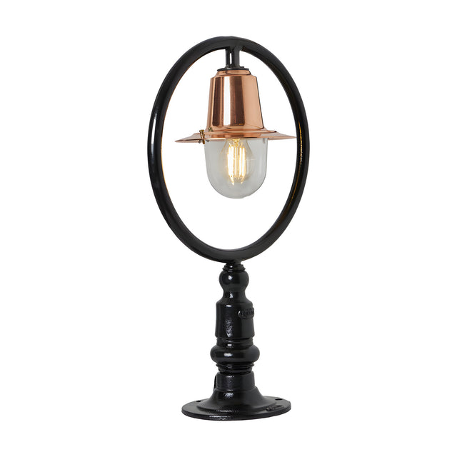 Small railway style pillar light with a black hoop frame, copper spun shade, and clear glass dome on a black cast pier base, plain white background.