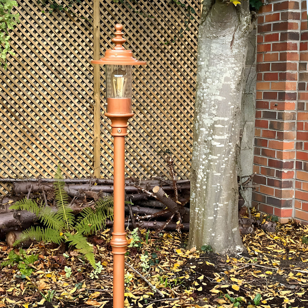 Georgian cast iron lamp post in copper-tone finish with lantern-style head and visible bulb, installed outdoors in a garden setting against a trellis fence and tree.
