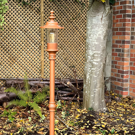 Georgian cast iron lamp post in copper-tone finish with lantern-style head and visible bulb, installed outdoors in a garden setting against a trellis fence and tree.