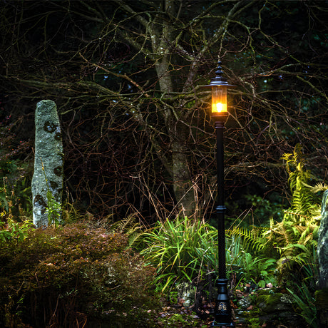 Georgian Cast Iron Lamp Post 2.21m in black, single lantern head with amber glow, mounted on a slim traditional pole in a garden setting amid ferns and a stone marker.