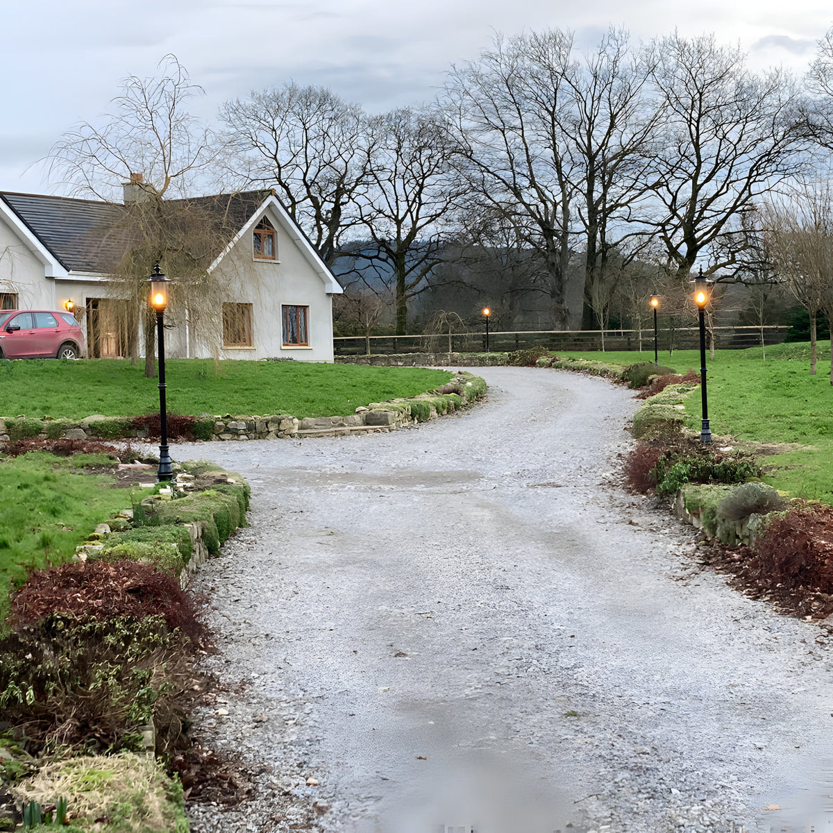 Georgian Cast Iron Lamp Post 2.21m in black lining a gravel driveway, traditional square design with shallow crown, lit outdoors beside a garden and cottage.