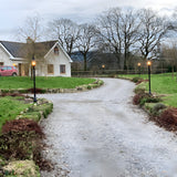 Georgian Cast Iron Lamp Post 2.21m in black lining a gravel driveway, traditional square design with shallow crown, lit outdoors beside a garden and cottage.