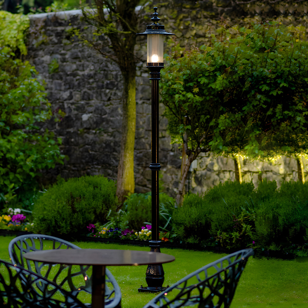 Georgian Cast Iron Lamp Post 2.21m in black, traditional post light with lantern head and glass panels illuminated on a lawn beside a garden table and stone wall.