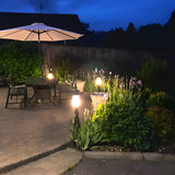 Large Georgian cast iron pedestal post lights in black, illuminated on a garden patio edge beside plants and outdoor seating at dusk.