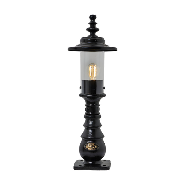 Small Georgian cast iron pedestal post light in black with clear cylindrical glass, decorative crown finial, and square mounting base, shown lit on a plain white background.
