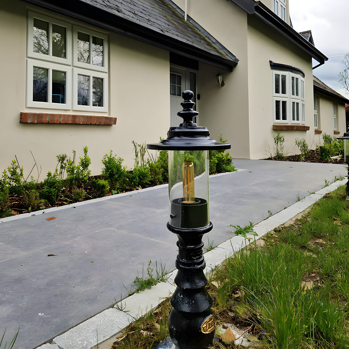 Small Georgian cast iron pedestal post light in black with clear cylindrical shade and visible bulb, shown on a driveway edge outside a period-style house.
