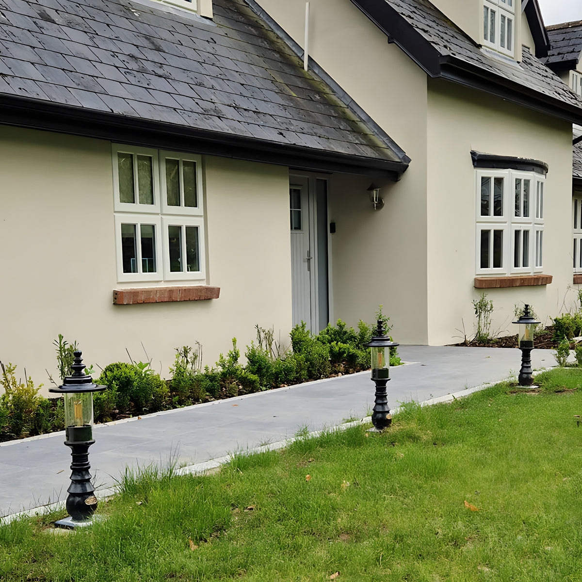 Small Georgian cast iron pedestal post lights in black lining a garden pathway leading to a house entrance, each with a square base and lantern-style glass head.