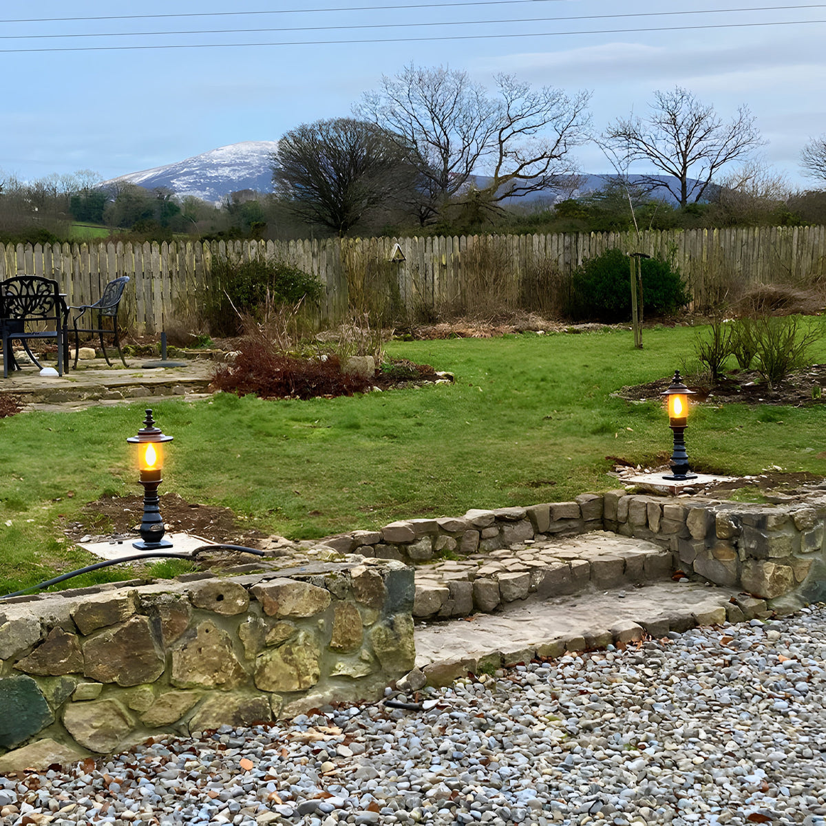 Small Georgian cast iron pedestal post light in black, lit with an amber glow, mounted on a garden lawn edge beside a stone wall and steps.