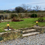 Small Georgian cast iron pedestal post light in black, lit with an amber glow, mounted on a garden lawn edge beside a stone wall and steps.