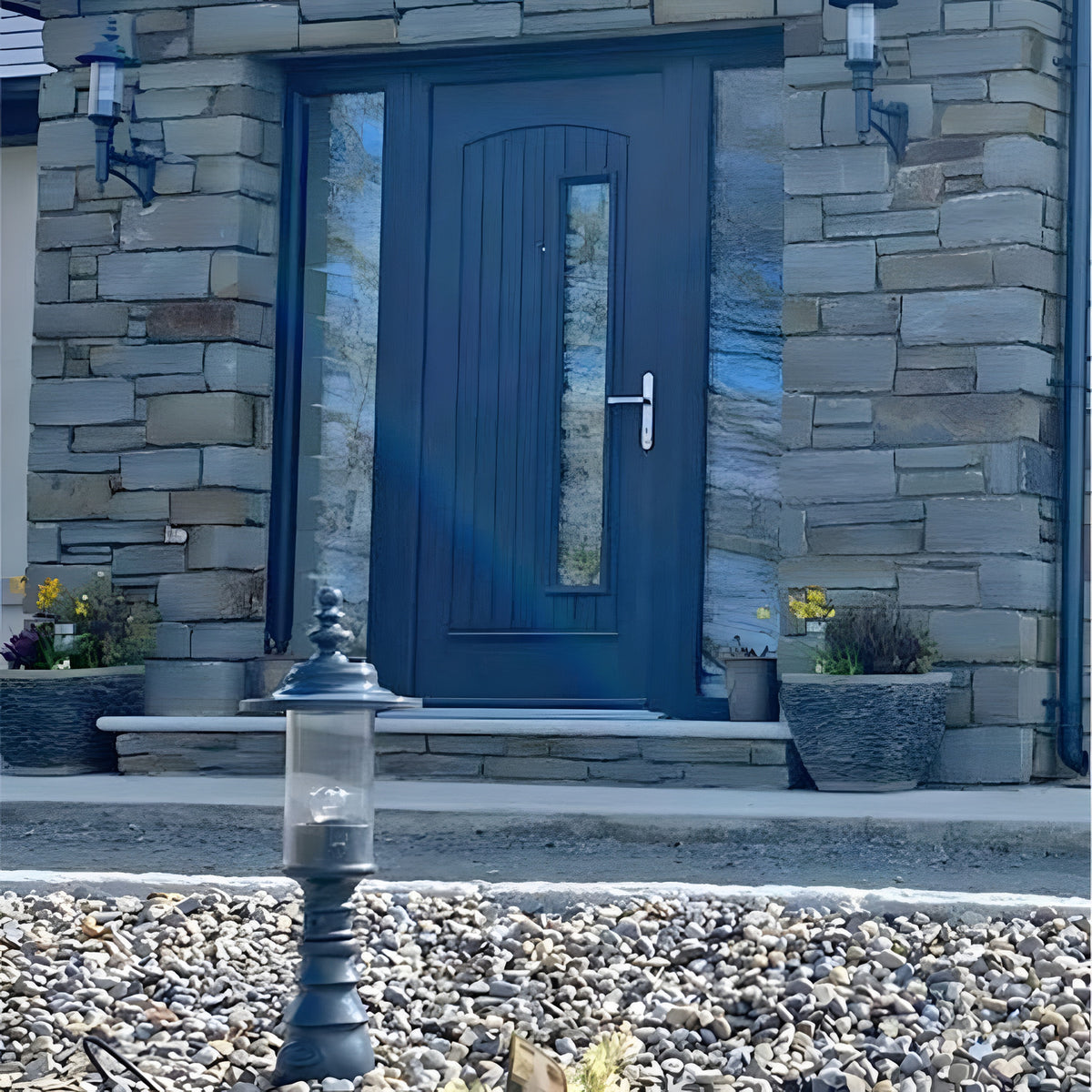Small Georgian cast iron pedestal post light in black with clear polycarbonate lantern, shown on gravel outside a stone house with a dark front door.