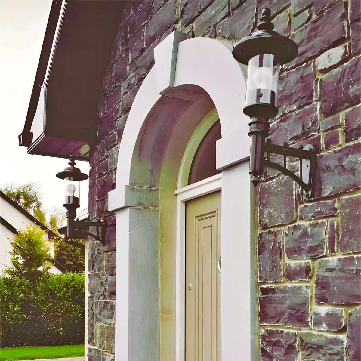Medium Georgian Cast Iron Wall Lantern in black mounted beside an arched stone doorway, showing its cylindrical glass shade, curved bracket and crown top on a stone house exterior.