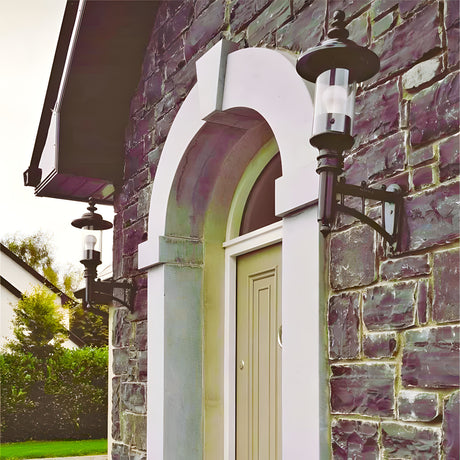 Medium Georgian Cast Iron Wall Lantern in black mounted beside an arched stone doorway, showing its cylindrical glass shade, curved bracket and crown top on a stone house exterior.