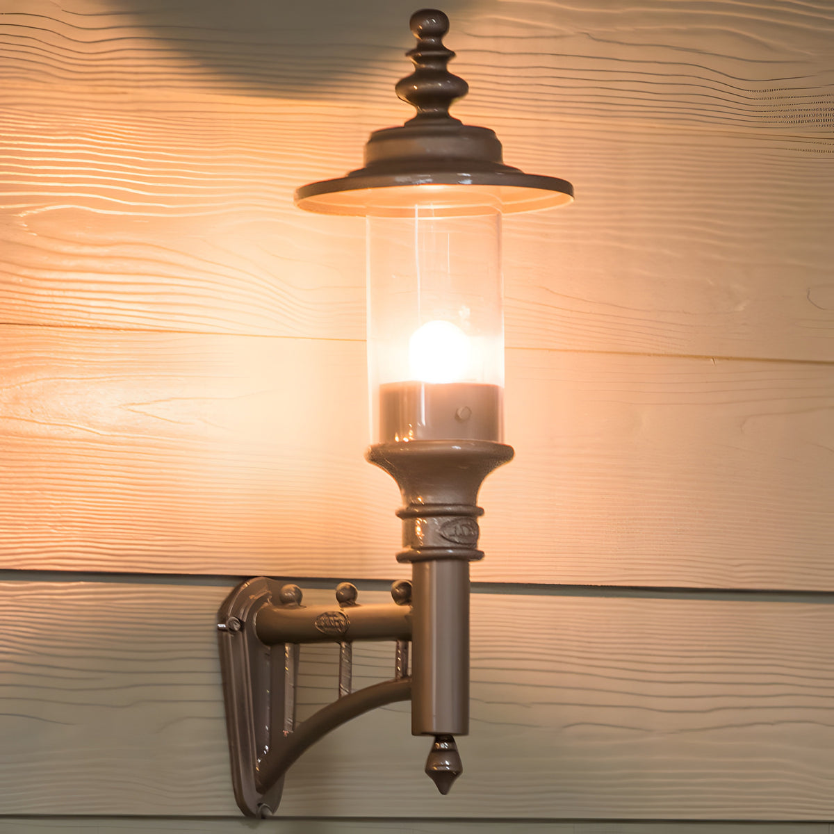 Small Georgian cast iron wall lantern in black, mounted on a wooden wall bracket with a lit bulb visible through the clear glass cylinder, set against wood panelling.