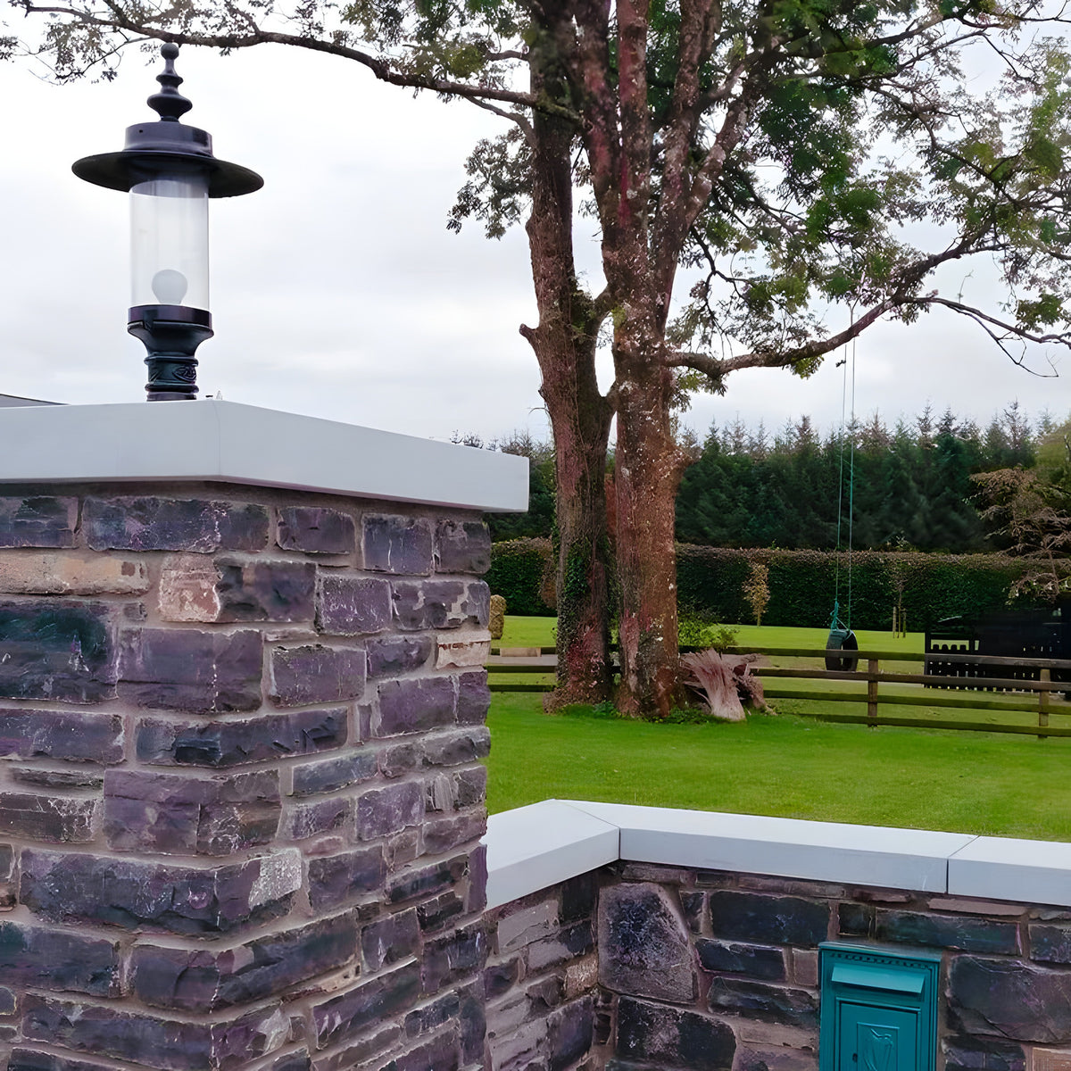 Large Georgian cast iron pillar light in black, with clear cylindrical shade, mounted on a stone pier cap at a garden entrance, trees and lawn visible beyond.