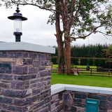 Large Georgian cast iron pillar light in black, with clear cylindrical shade, mounted on a stone pier cap at a garden entrance, trees and lawn visible beyond.