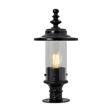 Small Georgian cast iron pillar light in black with clear cylindrical glass shade, ornate finial top and pier cap base, shown lit against a plain white background.