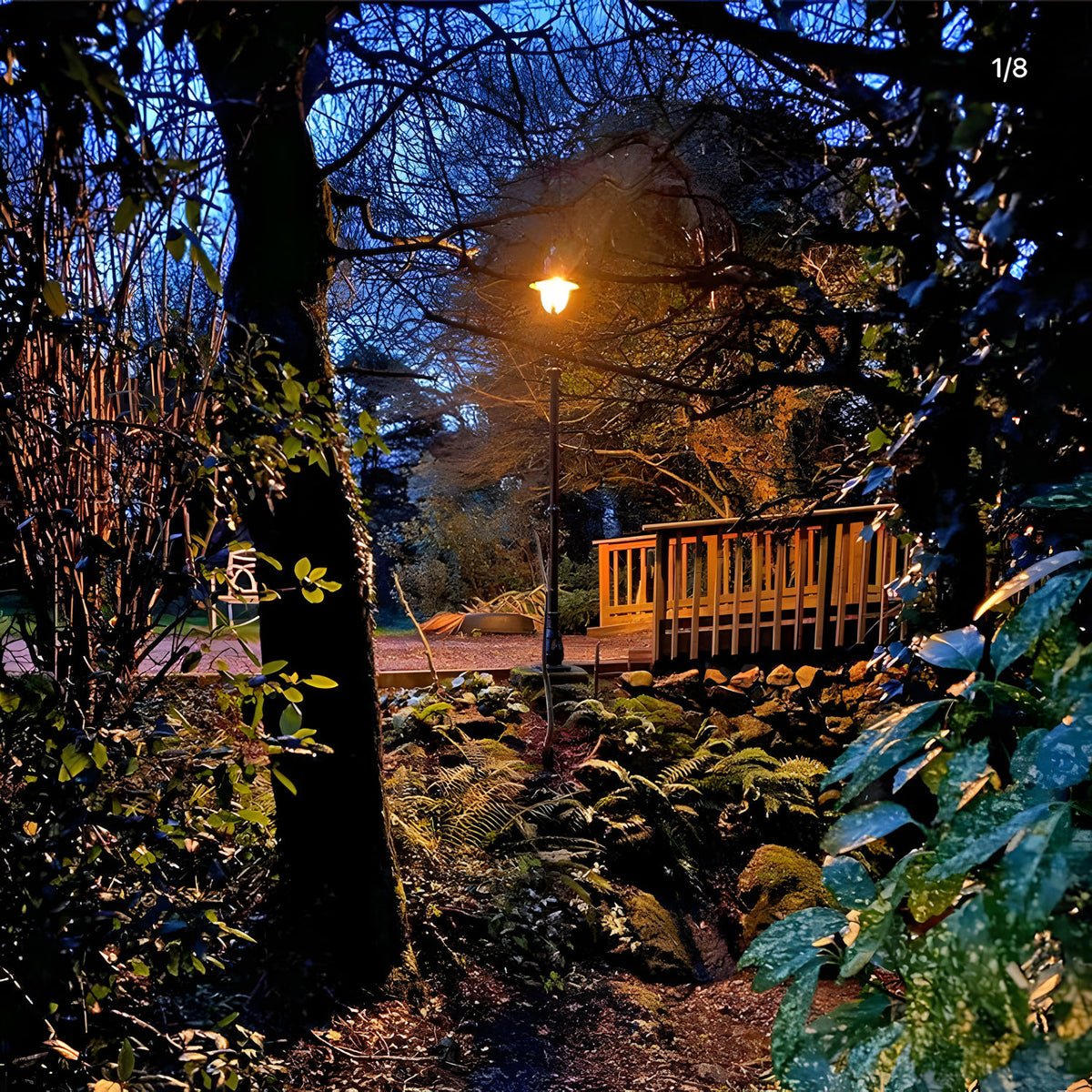 Vintage Cast Iron Lamp Post 2.8m in black, illuminated at dusk in a garden setting beside a wooden deck, ferns, and trees.