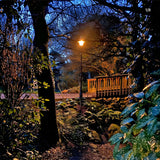 Vintage Cast Iron Lamp Post 2.8m in black, illuminated at dusk in a garden setting beside a wooden deck, ferns, and trees.