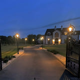 Vintage cast iron lamp posts, 2.5m black gooseneck design with teardrop shades, lighting a driveway leading to a house at dusk.