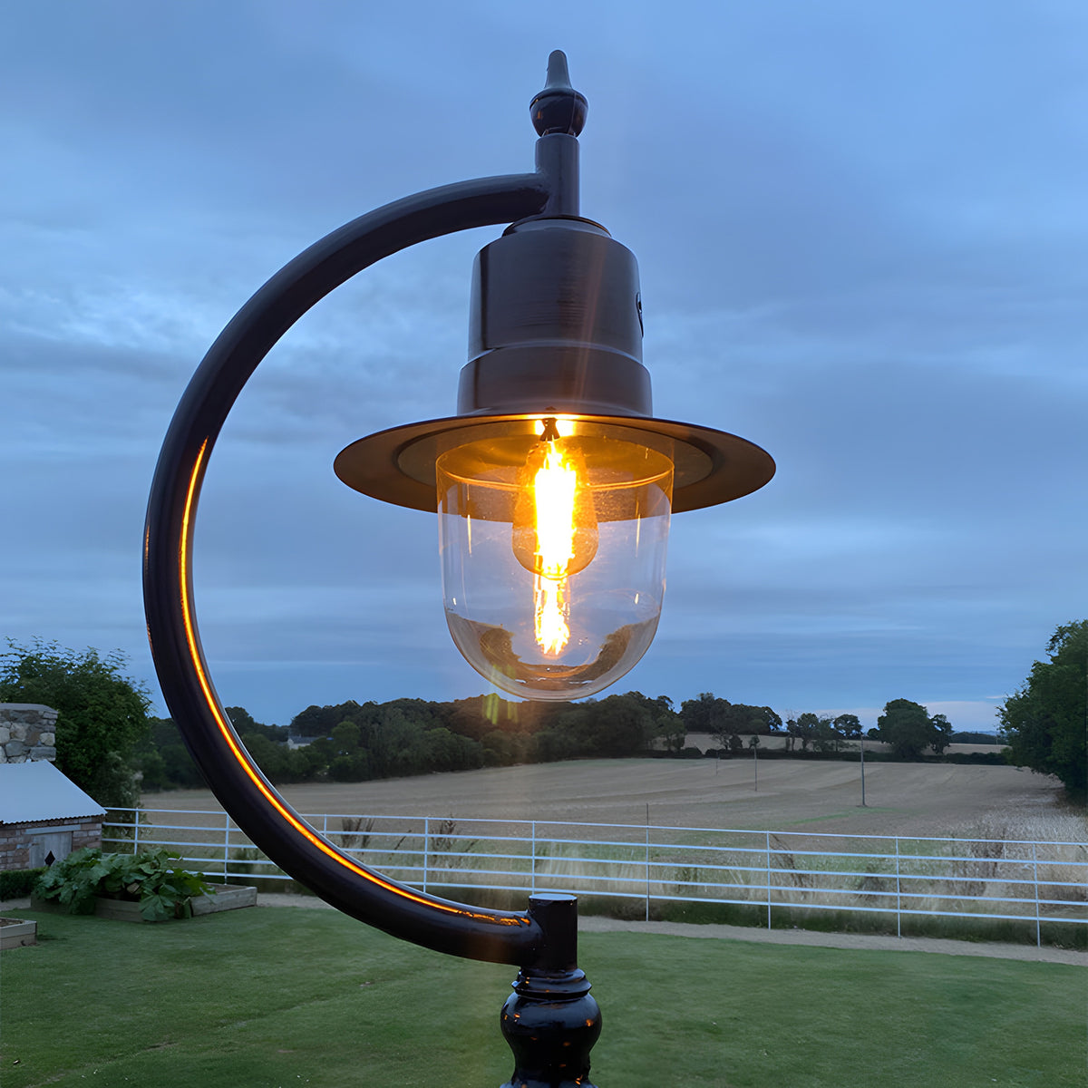 Vintage Cast Iron Lamp Post 2.5m in black with curved gooseneck arm, teardrop shade and glass dome lit at dusk, set against a garden and countryside backdrop.