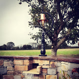 Large Vintage Cast Iron Pedestal Post Light with copper teardrop shade and gooseneck arm, mounted on a stone wall against a garden and tree backdrop.