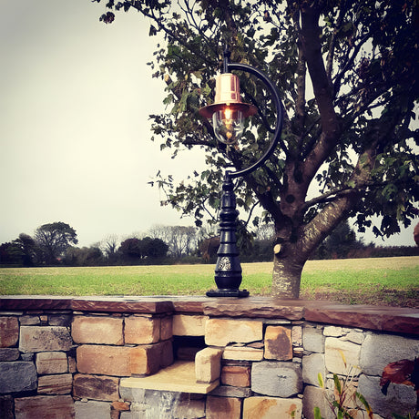 Large Vintage Cast Iron Pedestal Post Light with copper teardrop shade and gooseneck arm, mounted on a stone wall against a garden and tree backdrop.